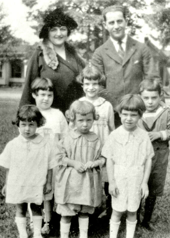 Photograph of six children, mostly dressed in white, in front of a man and a woman.